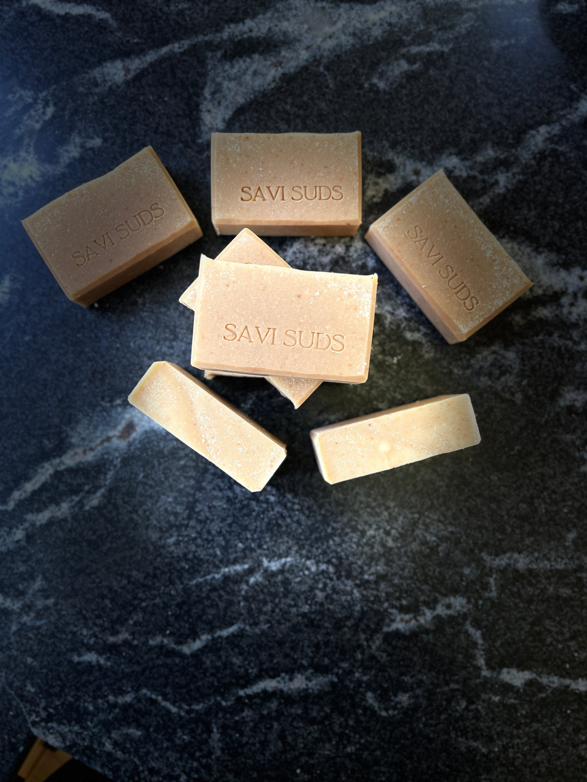Several rectangular bars of Savi Suds soap in warm beige and cream tones, arranged on a dark marbled surface. The brand name 'SAVI SUDS' is stamped on the top of the bars.