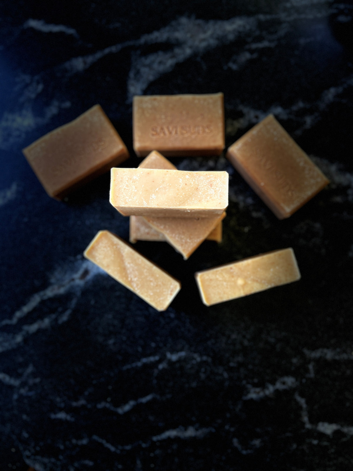 Several rectangular bars of soap in warm tan and light brown shades are arranged on a dark marbled surface. One bar in the center is stacked on top of others, and the soap bars show a slightly textured, natural finish.