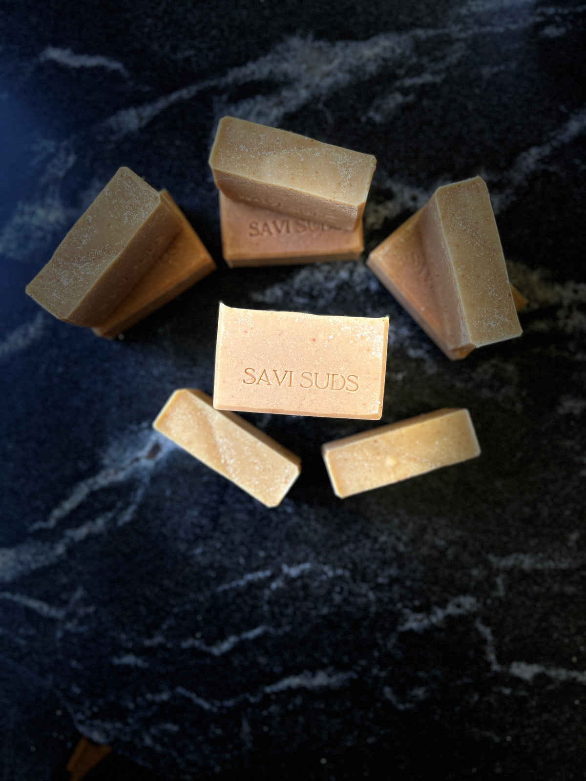 Nine rectangular bars of Savi Suds soap arranged on a dark, textured marble surface, with one bar centered in the foreground showing the brand name 'SAVI SUDS' stamped on the front.