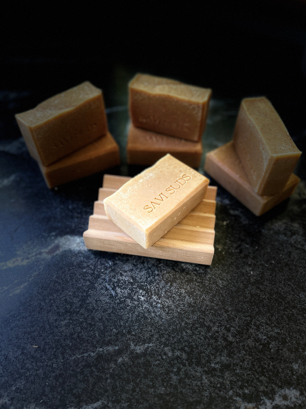 Several rectangular bars of soap in warm tan and brown tones are arranged on a dark textured surface, with bar in the foreground resting on a small wooden stand. The word SAVISUDS is stamped on the bars