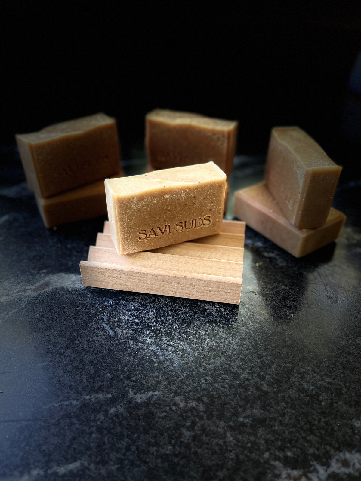Several rectangular bars of soap in warm tan and medium brown tones are arranged on a dark stone-like surface. One front bar is light tan and embossed with the brand name 'SAVI SUDS'.