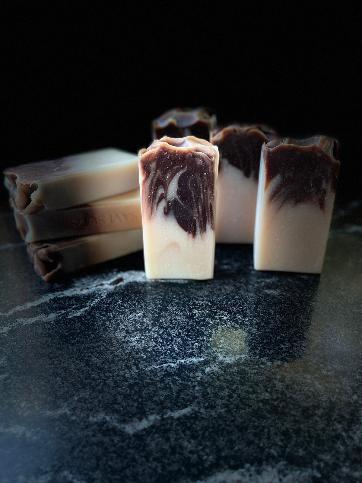 Several handmade rectangular bars of soap with a marbled cream and dark brown swirl pattern, arranged on a dark stone surface against a black background.