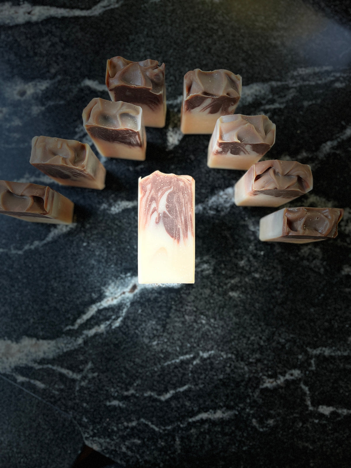 A handmade soap bar with a cream and dark brown marbled swirl pattern, standing vertically in the center of a dark marble surface, surrounded by eight identical soap bars arranged in a circle.