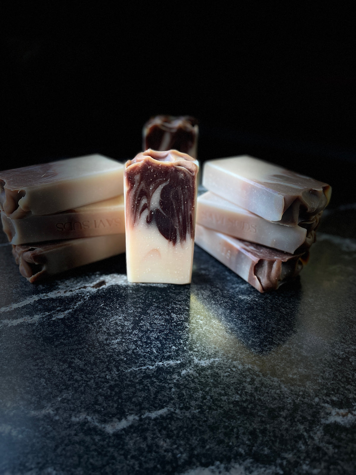 Several handmade rectangular bars of soap with a marbled cream and dark brown swirl pattern, arranged on a dark, textured surface against a black background.