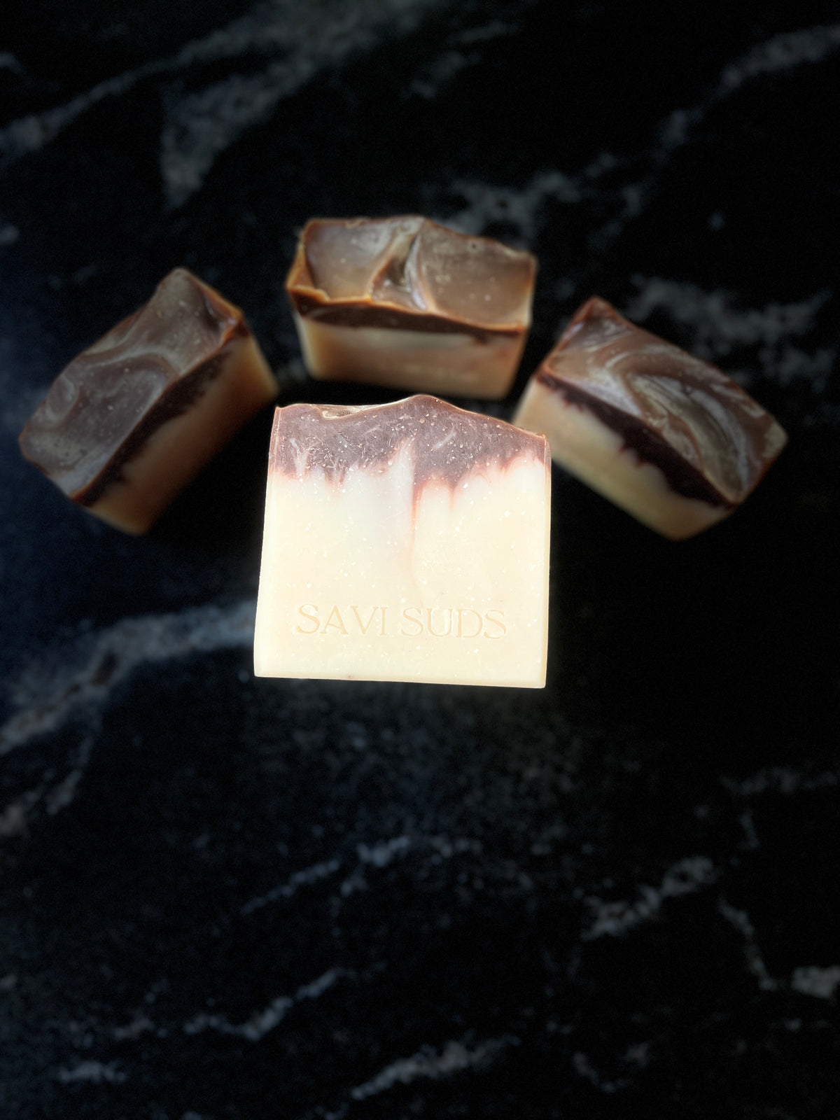 Four square bars of handmade soap on a black marble surface, with one bar in the foreground stamped with the brand name 'SAVI SUDS'. The soap features a marbled pattern of cream, dark brown, and lighter beige tones.