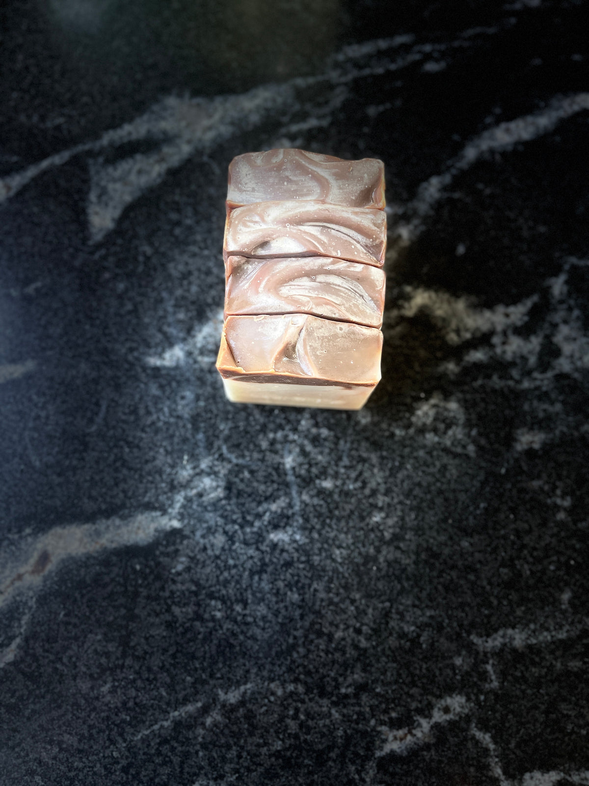 Four square bars of handmade soap lined up in a row with a top view of the swirled brown pattern, placed on a dark marble surface.