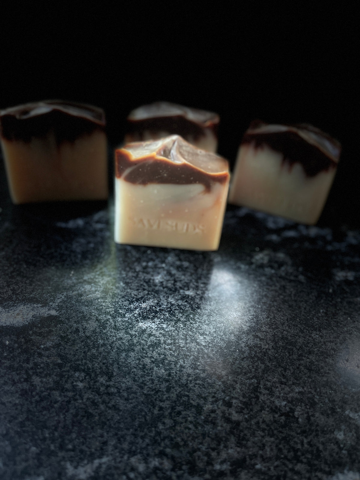 Four square bars of handmade soap with a cream and dark brown marbled pattern, arranged on a dark textured surface against a black background.