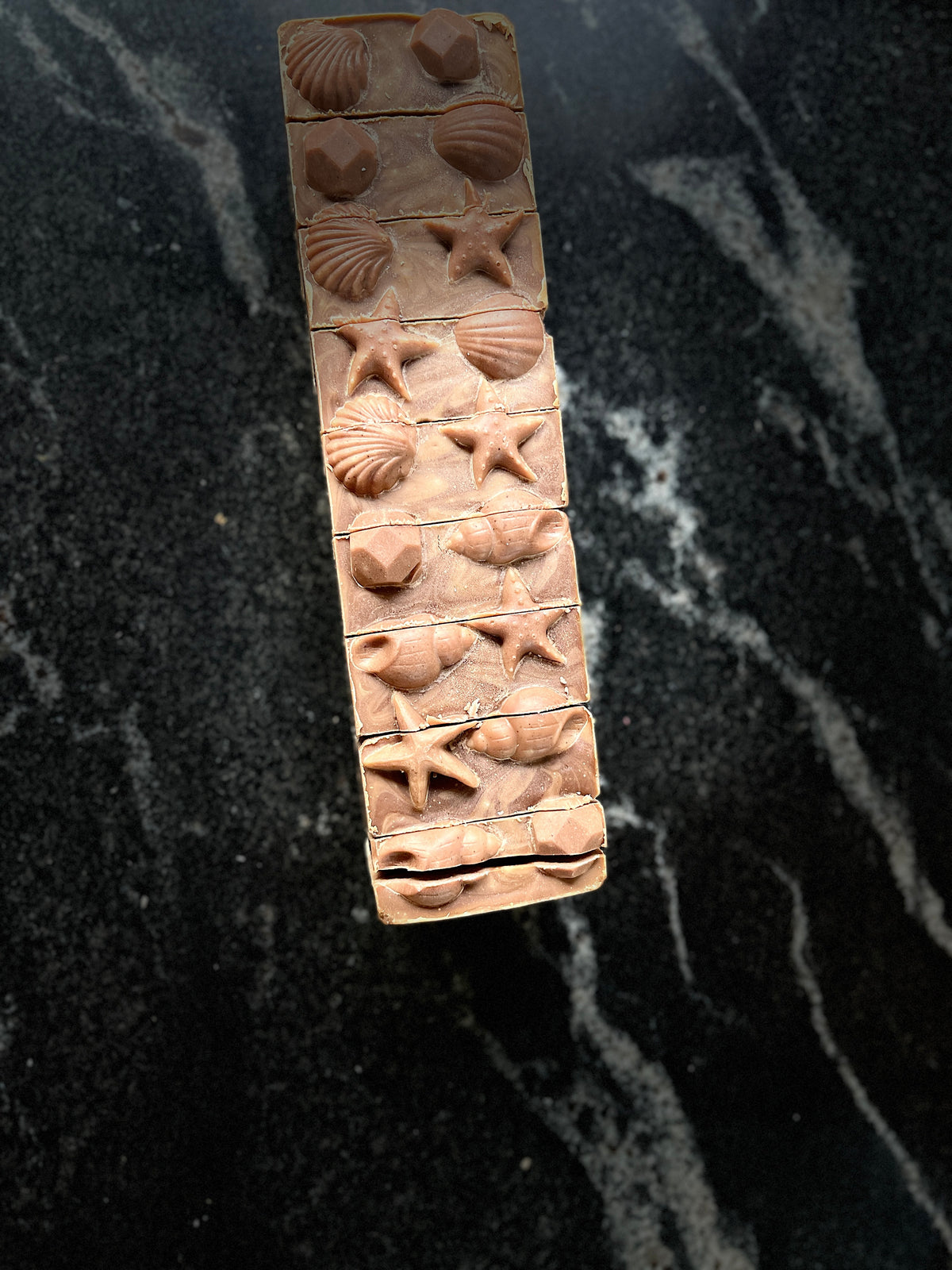 A rectangular bar of handmade soap with a brown top layer featuring brown seashells and star motifs, displayed on a dark marble surface.