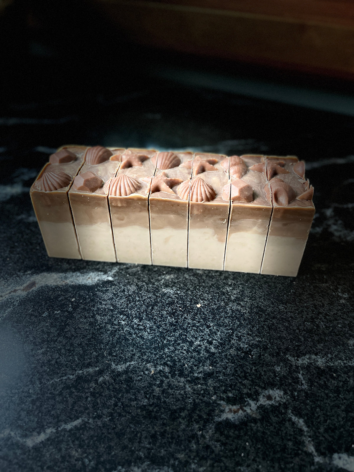 A rectangular bar of layered soap with a beige base and a top layer featuring brown seashell and star motifs, displayed on a dark marble surface.