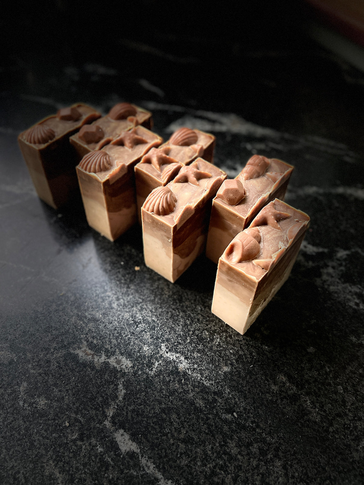 A side view of 7 warm vanilla soaps lined up on a dark marbled surface. Soaps are topped with sea shells and star motifs