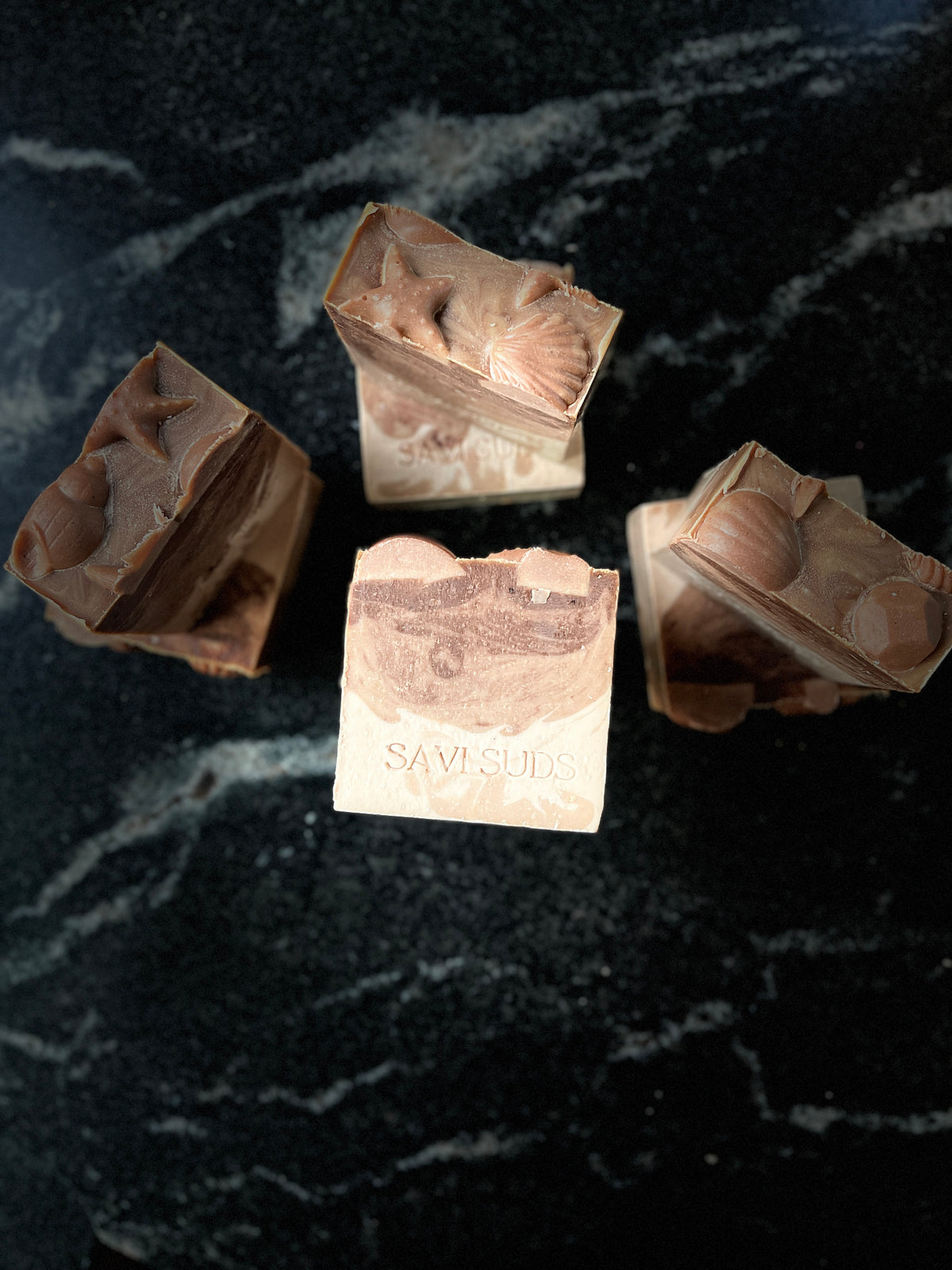 Seven warm vanilla soaps arranged on a dark marbled surface. The middle soap is elevated to show detail and SAVI SUDS brand name is stamped on the soap