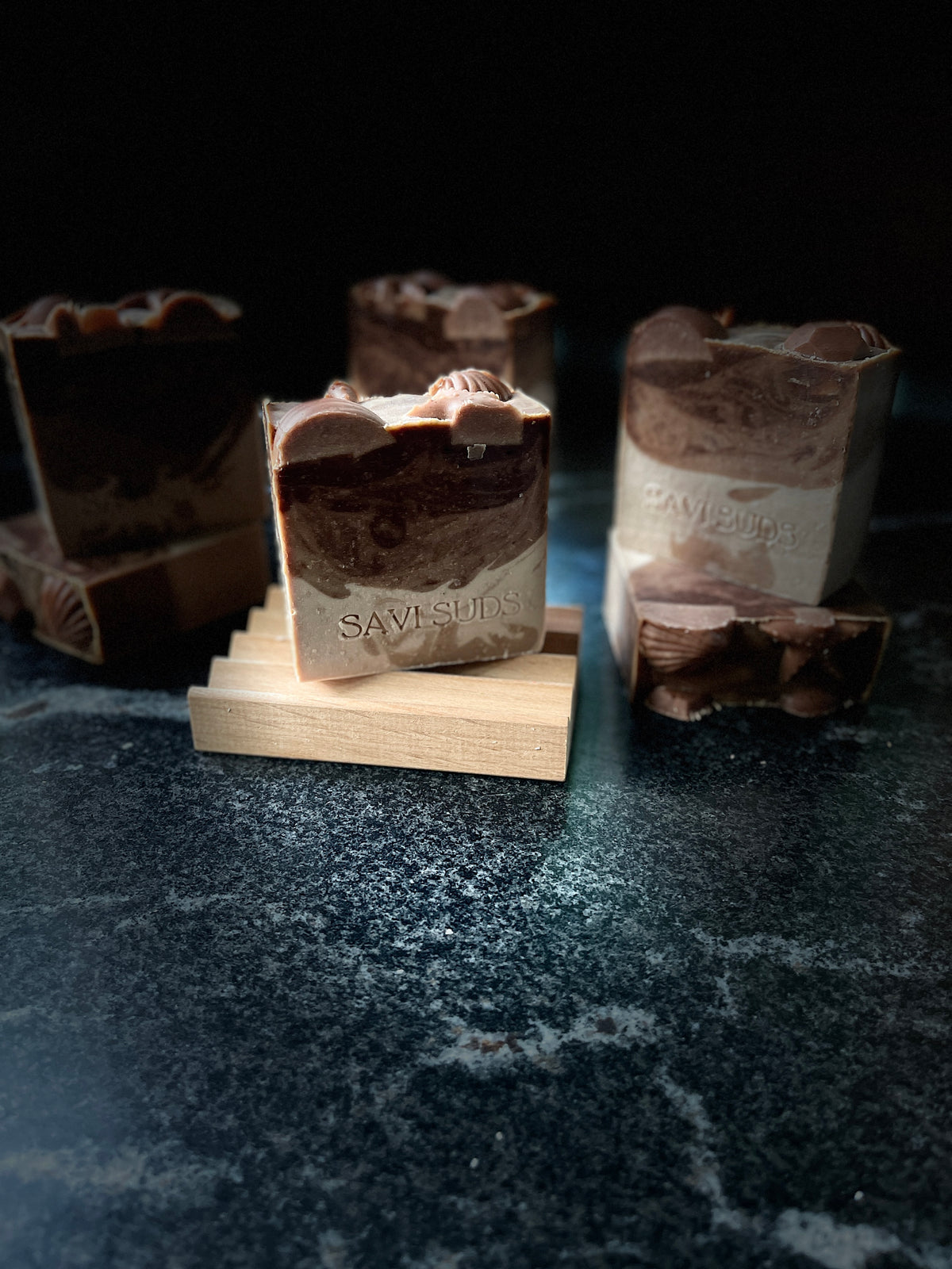 A warm vanilla soap is resting on a wooden soap dish with other warm vanilla soaps blurred in the background.  SAVI SUDS is stamped on the front of each bar