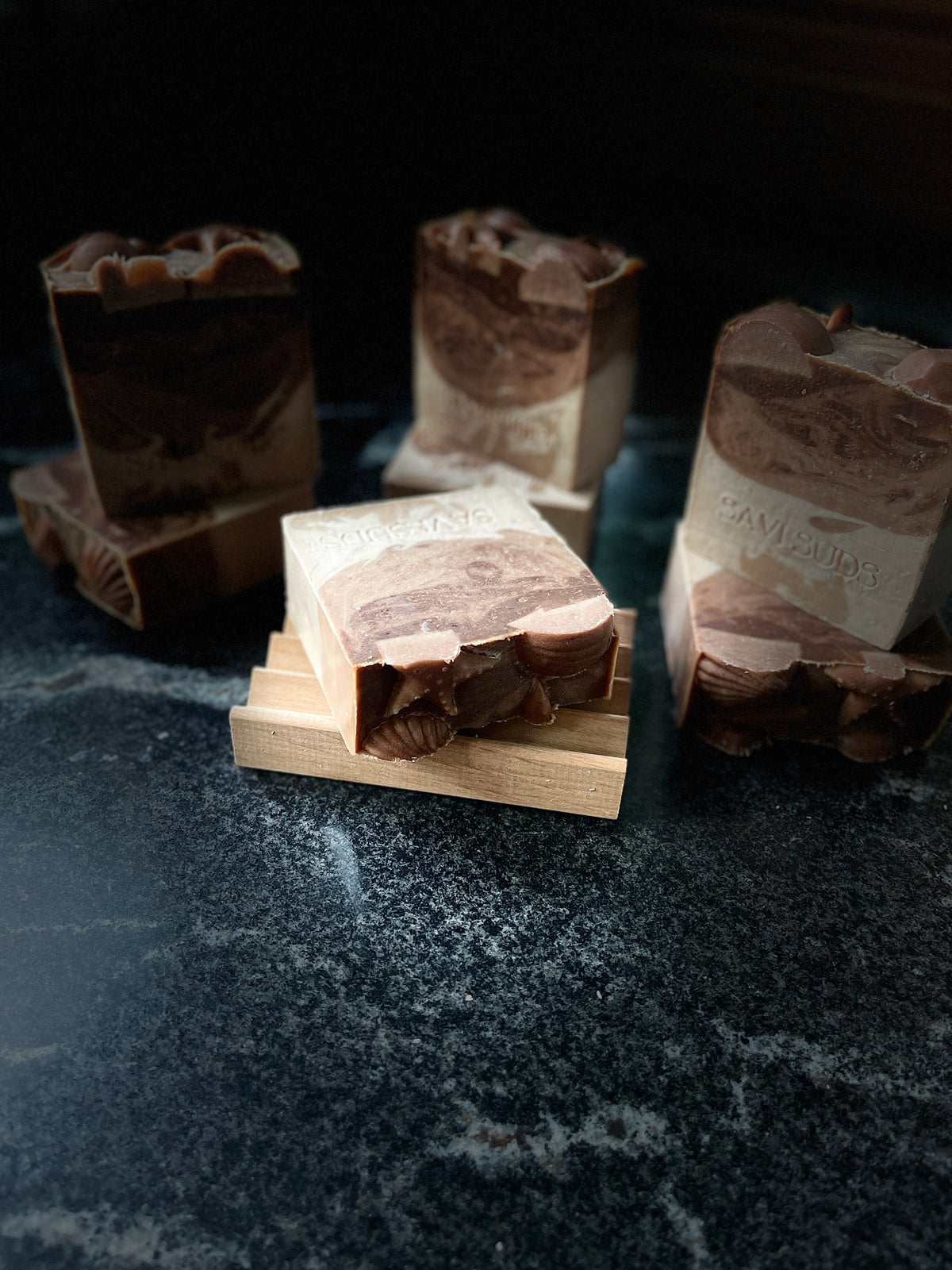 Seven warm vanilla soaps are arranged on a dark marbled surface with one in the forefront resting on a wooden soap dish. SAVI SUDS is stamped on each soap front
