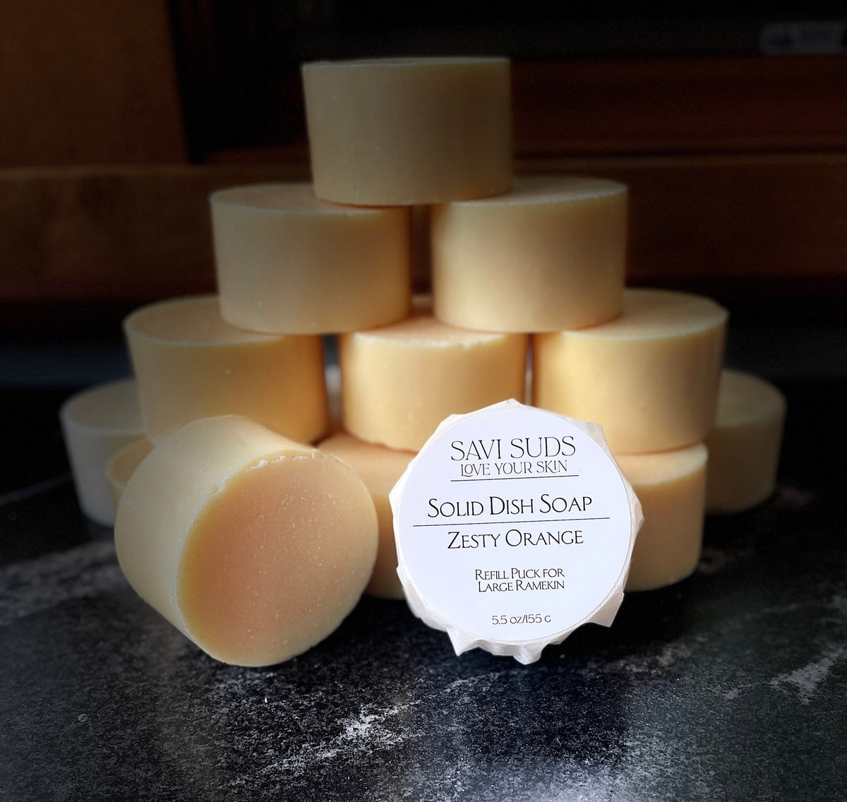A stack of solid dish soap refill bars with one in front wrapped in bleach free coffee filter that reads 'SAVI SUDS' and 'SOLID DISH SOAP ZESTY ORANGE', set on a dark textured countertop.