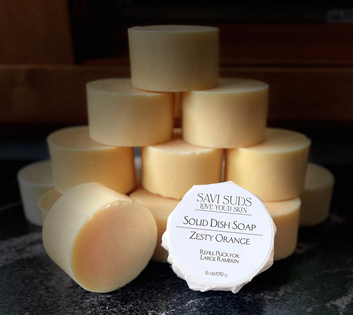 A stack of solid dish soap refill bars with one in front wrapped in bleach free coffee filter that reads 'SAVI SUDS' and 'SOLID DISH SOAP ZESTY ORANGE', set on a dark textured countertop.