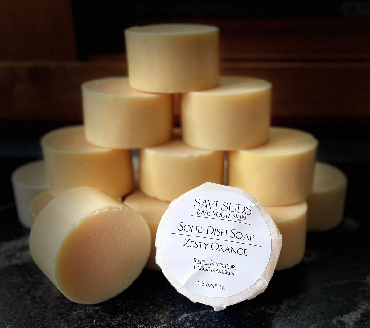 A stack of solid dish soap refill bars with one in front wrapped in bleach free coffee filter that reads 'SAVI SUDS' and 'SOLID DISH SOAP ZESTY ORANGE', set on a dark textured countertop.