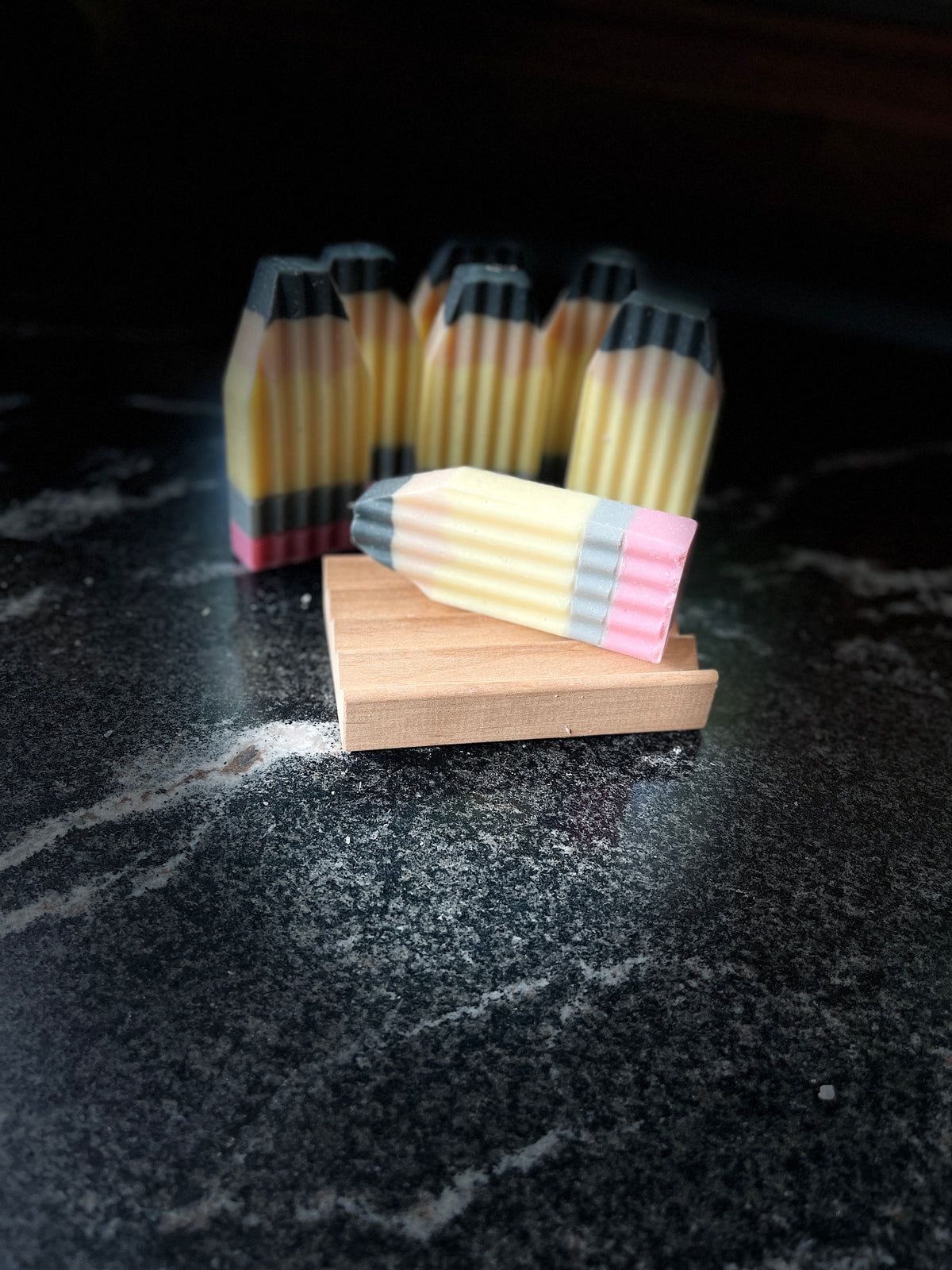 A set of novelty pencil-shaped soaps displayed in the background with one soap in sharp focus on a small wooden block in the foreground