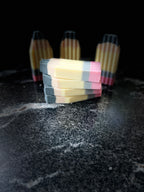 Four rectangular handmade soaps shaped like pencils, stacked in the foreground with three standing upright in the background on a dark textured surface.