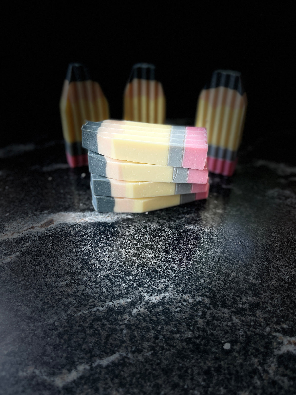 Four rectangular handmade soaps shaped like pencils, stacked in the foreground with three standing upright in the background on a dark textured surface.